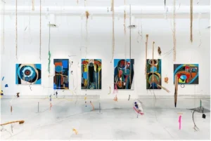 An art installation by Merikokeb Berhanu at the 2022 Venice Biennale, featuring large abstract blue and earth-toned paintings on white walls with organic fibers hanging from the ceiling.