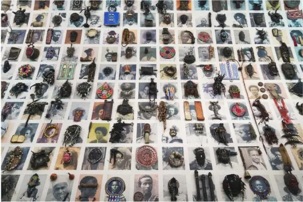 A large-scale mixed-media installation by Wendimagegn Belete titled Your Gaze Makes Me, featuring a grid of numerous portraits overlaid with diverse physical objects, masks, and cultural artifacts.